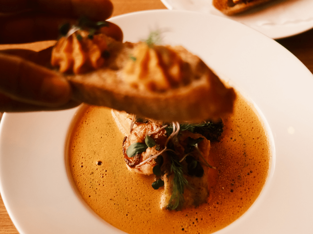 Jemand hält ein Knoblauchbaguette in die Kamera im Restaurant und Landhotel Winter. Dahinter zu sehen ein Teller der Bouillabaisse.